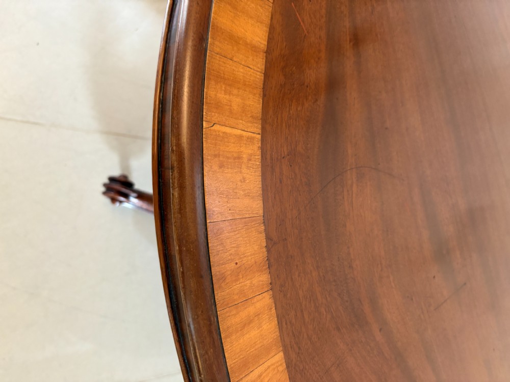 An antique Victorian oval mahogany dining table featuring crossbanded satinwood inlay, shown tilted vertically on a carved quadruped base with scroll feet and brass castors. View III
