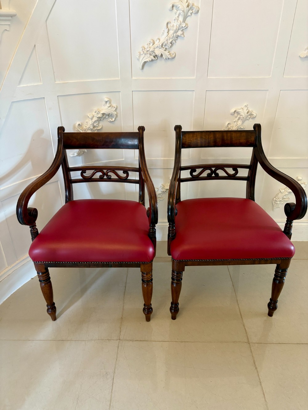 A pair of George III mahogany desk chairs featuring scroll arms, carved back splats, turned front legs, and red leather seats.