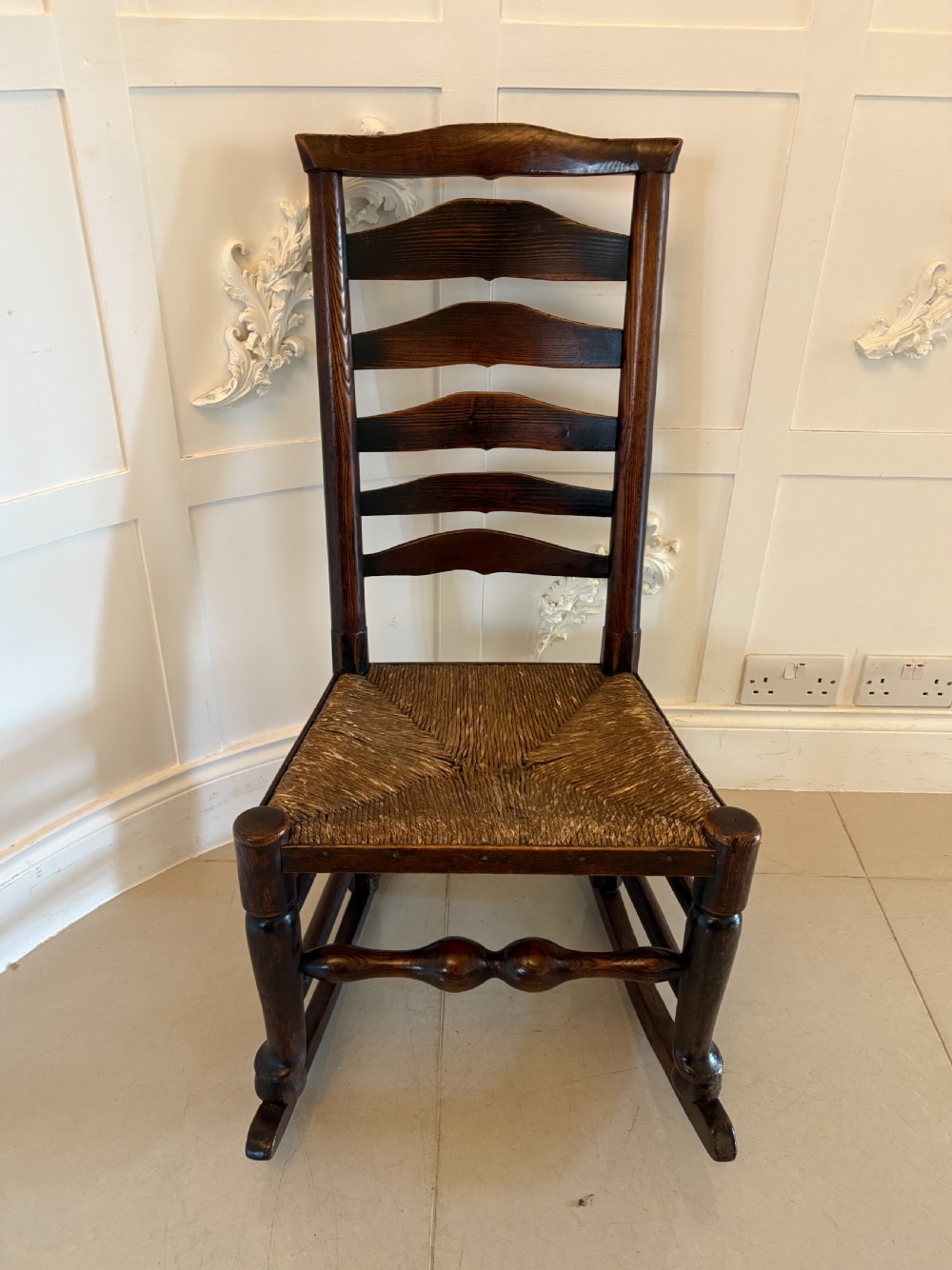 An 18th-century dark oak ladder back rocking chair featuring five shaped horizontal back slats, a woven rush seat, turned front legs, and wooden rockers.