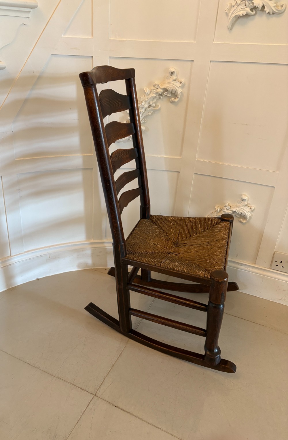 An 18th-century dark oak ladder back rocking chair featuring five shaped horizontal back slats, a woven rush seat, turned front legs, and wooden rockers. View III