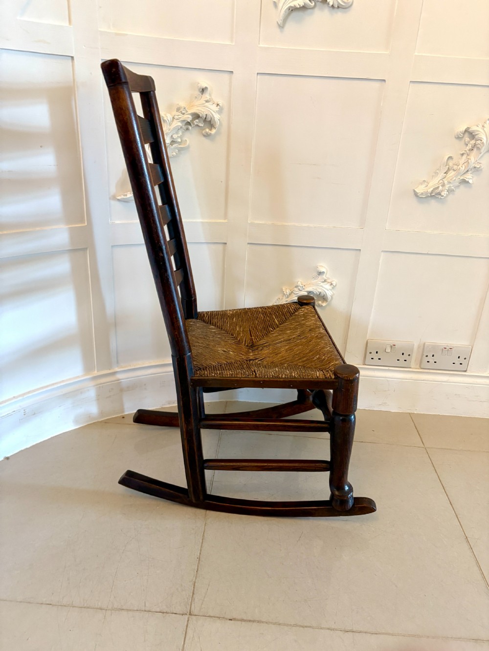 An 18th-century dark oak ladder back rocking chair featuring five shaped horizontal back slats, a woven rush seat, turned front legs, and wooden rockers. View V