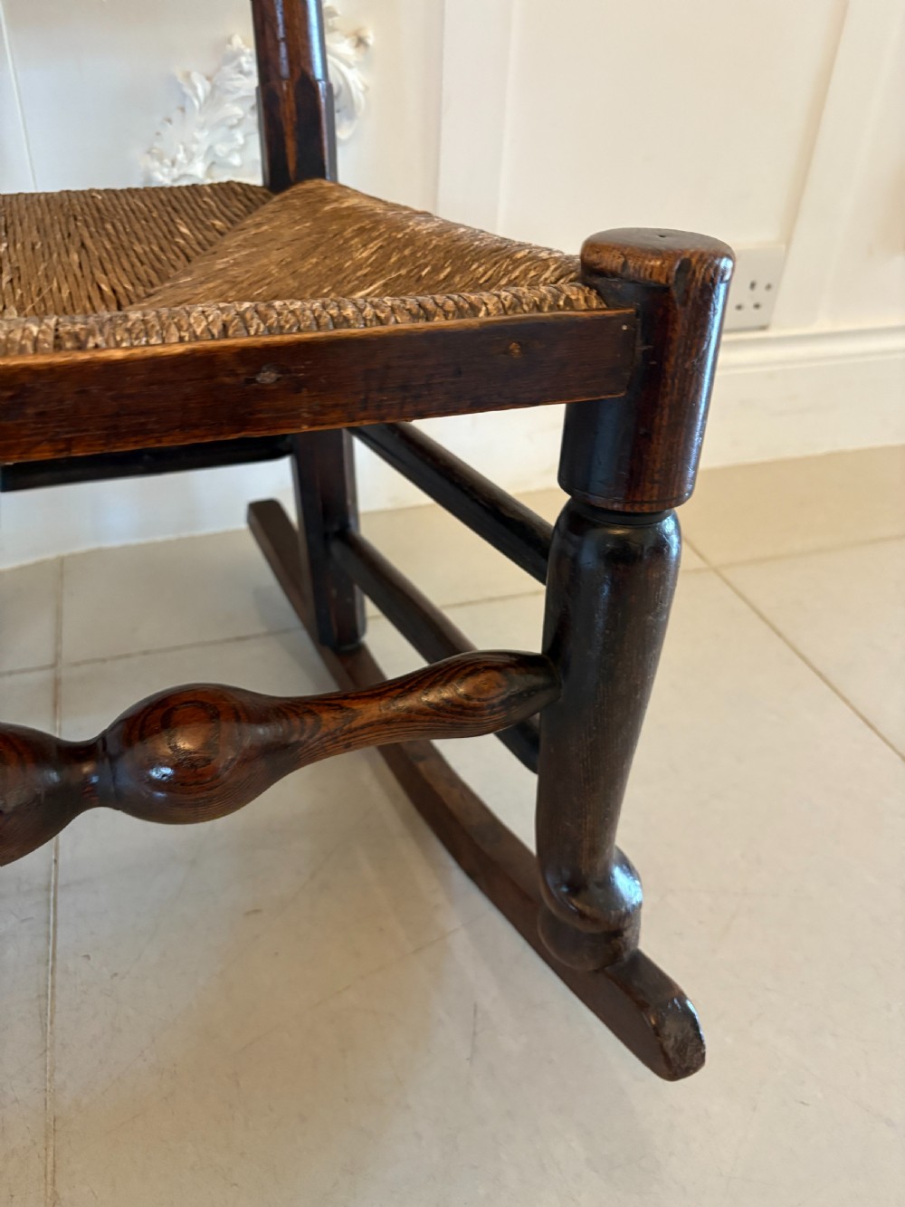 An 18th-century dark oak ladder back rocking chair featuring five shaped horizontal back slats, a woven rush seat, turned front legs, and wooden rockers. View VI