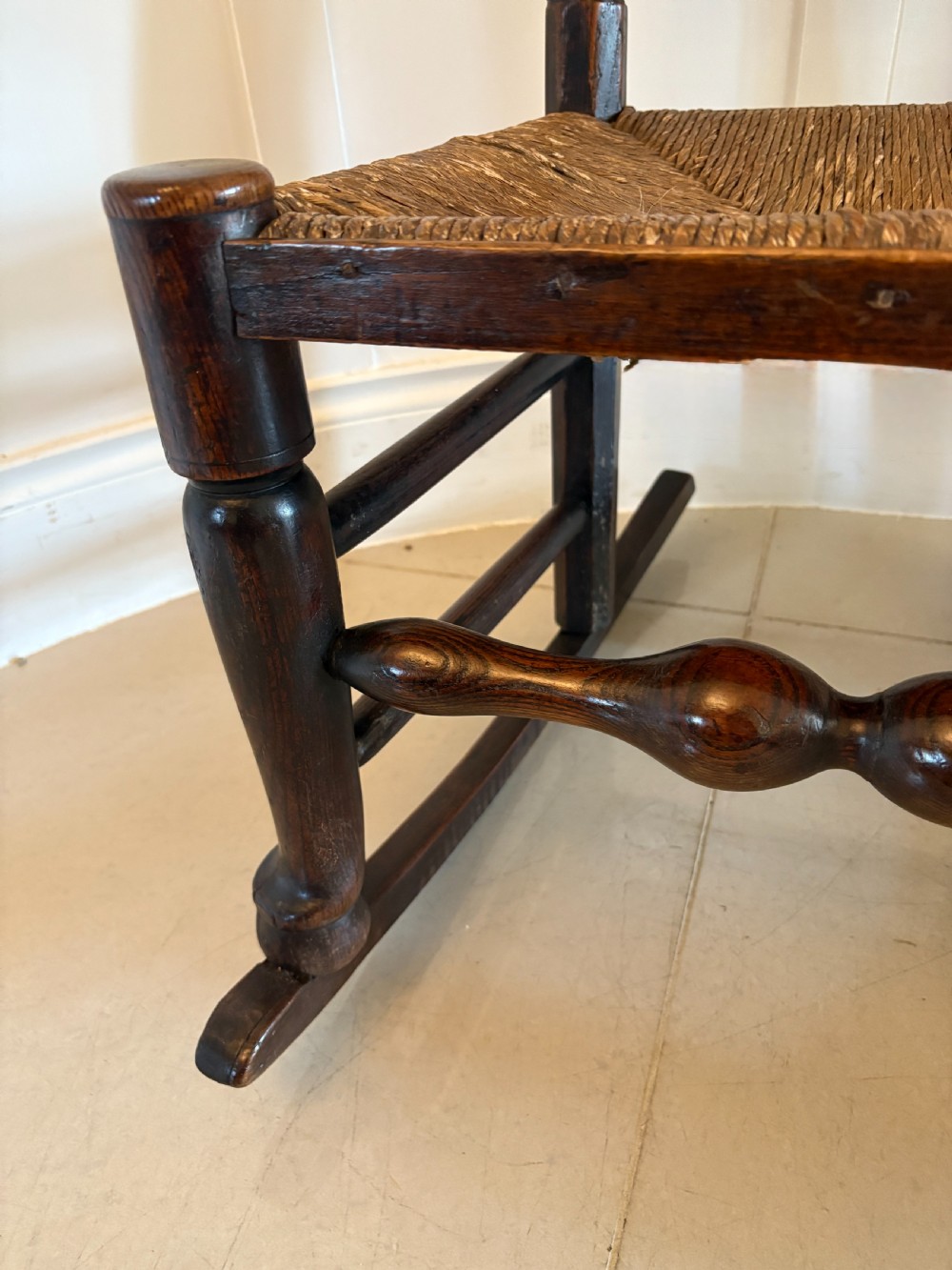 An 18th-century dark oak ladder back rocking chair featuring five shaped horizontal back slats, a woven rush seat, turned front legs, and wooden rockers. View VII