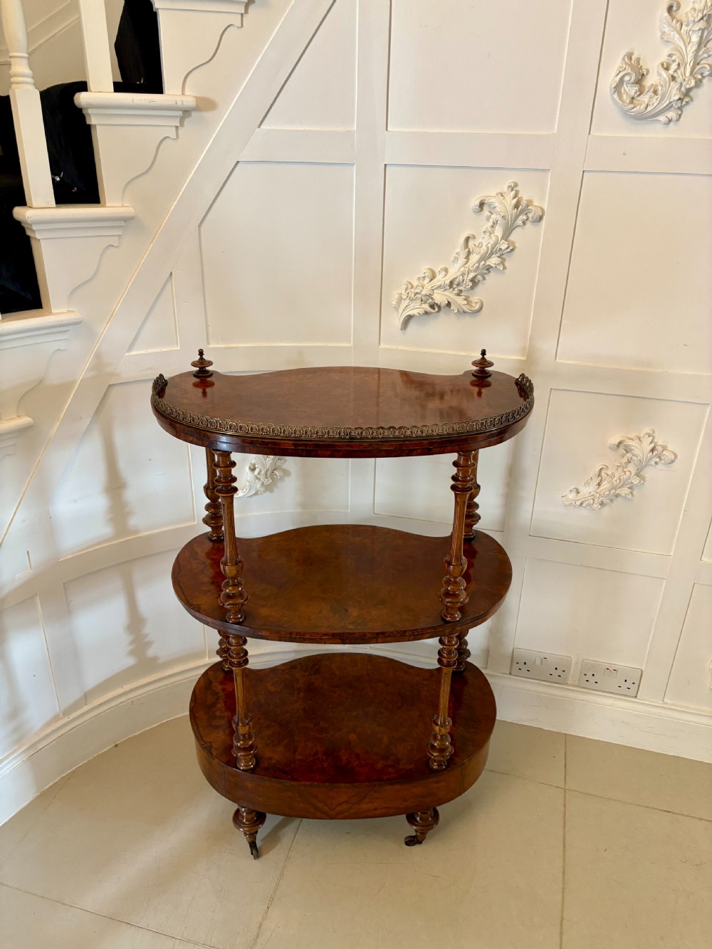 A Victorian three-tiered burr walnut whatnot featuring serpentine shelves, turned spindle supports, a pierced brass top gallery, and a base drawer on brass castors. View III