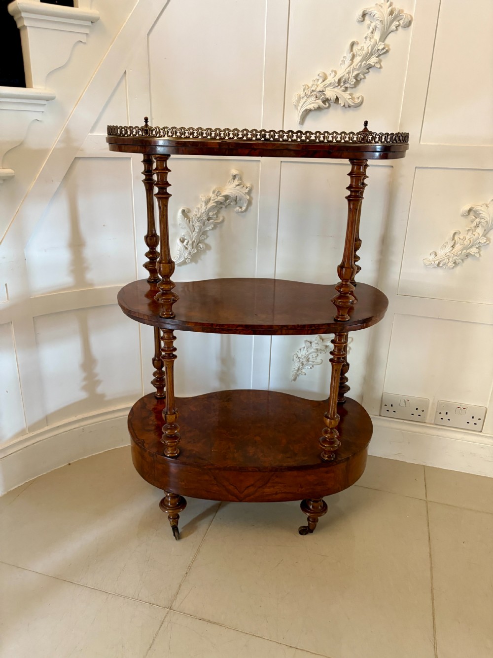 A Victorian three-tiered burr walnut whatnot featuring serpentine shelves, turned spindle supports, a pierced brass top gallery, and a base drawer on brass castors. View IV