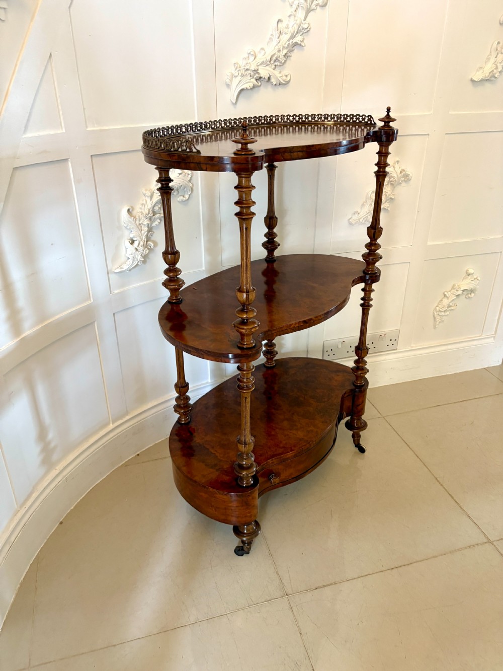 A Victorian three-tiered burr walnut whatnot featuring serpentine shelves, turned spindle supports, a pierced brass top gallery, and a base drawer on brass castors. View XII