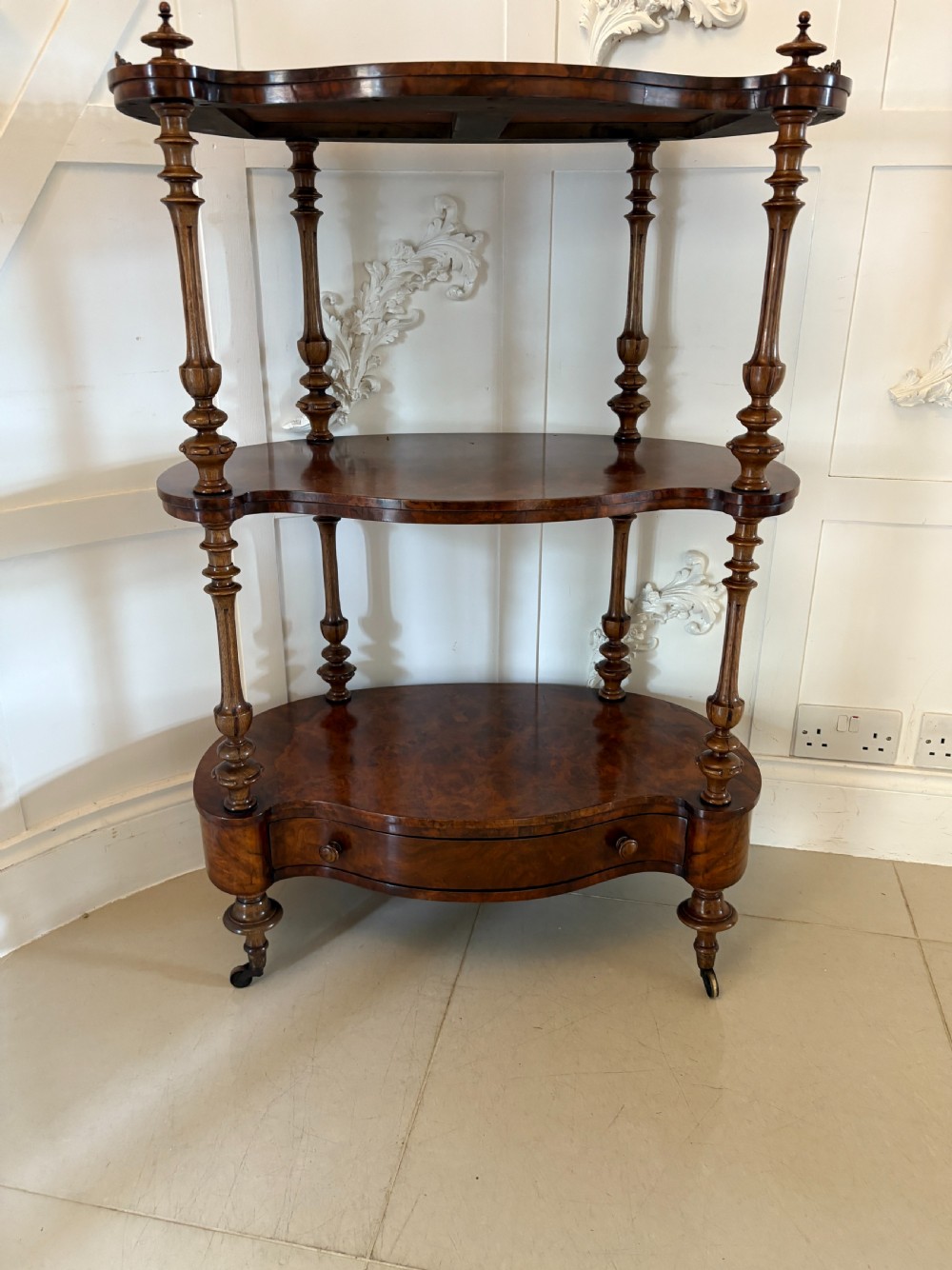 A Victorian three-tiered burr walnut whatnot featuring serpentine shelves, turned spindle supports, a pierced brass top gallery, and a base drawer on brass castors. View XVI