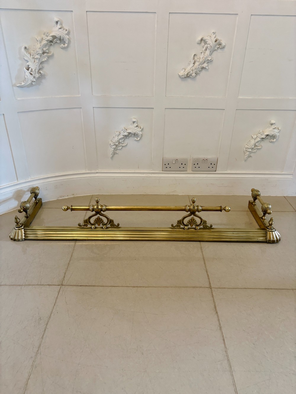A Victorian brass fireplace fender featuring a low moulded base, fluted corner finials, and an elevated tool rail supported by scrolling brackets.