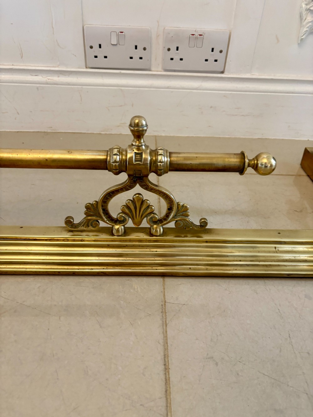 A Victorian brass fireplace fender featuring a low moulded base, fluted corner finials, and an elevated tool rail supported by scrolling brackets. View V