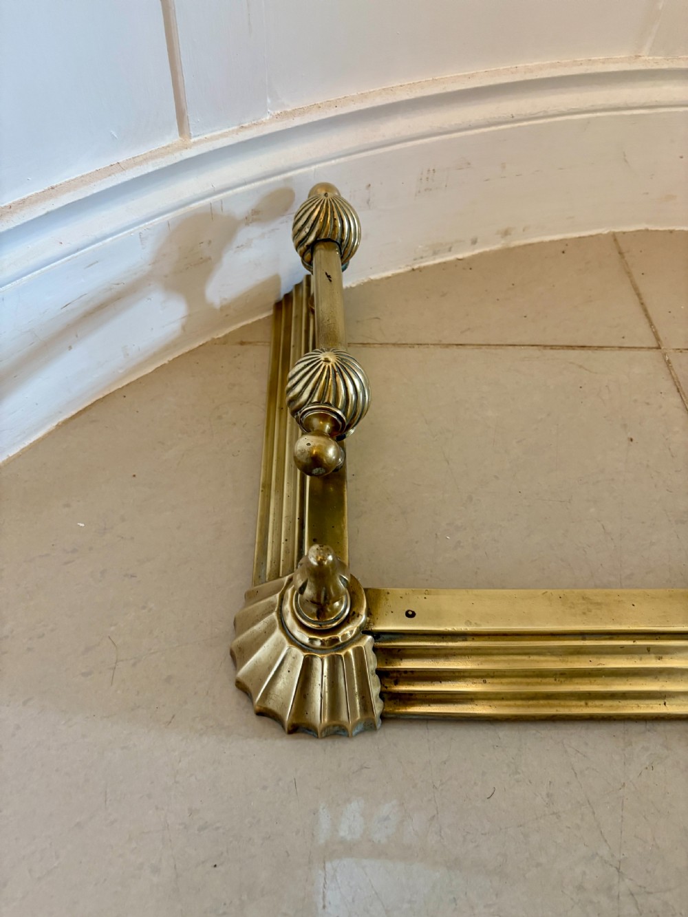 A Victorian brass fireplace fender featuring a low moulded base, fluted corner finials, and an elevated tool rail supported by scrolling brackets. View VI