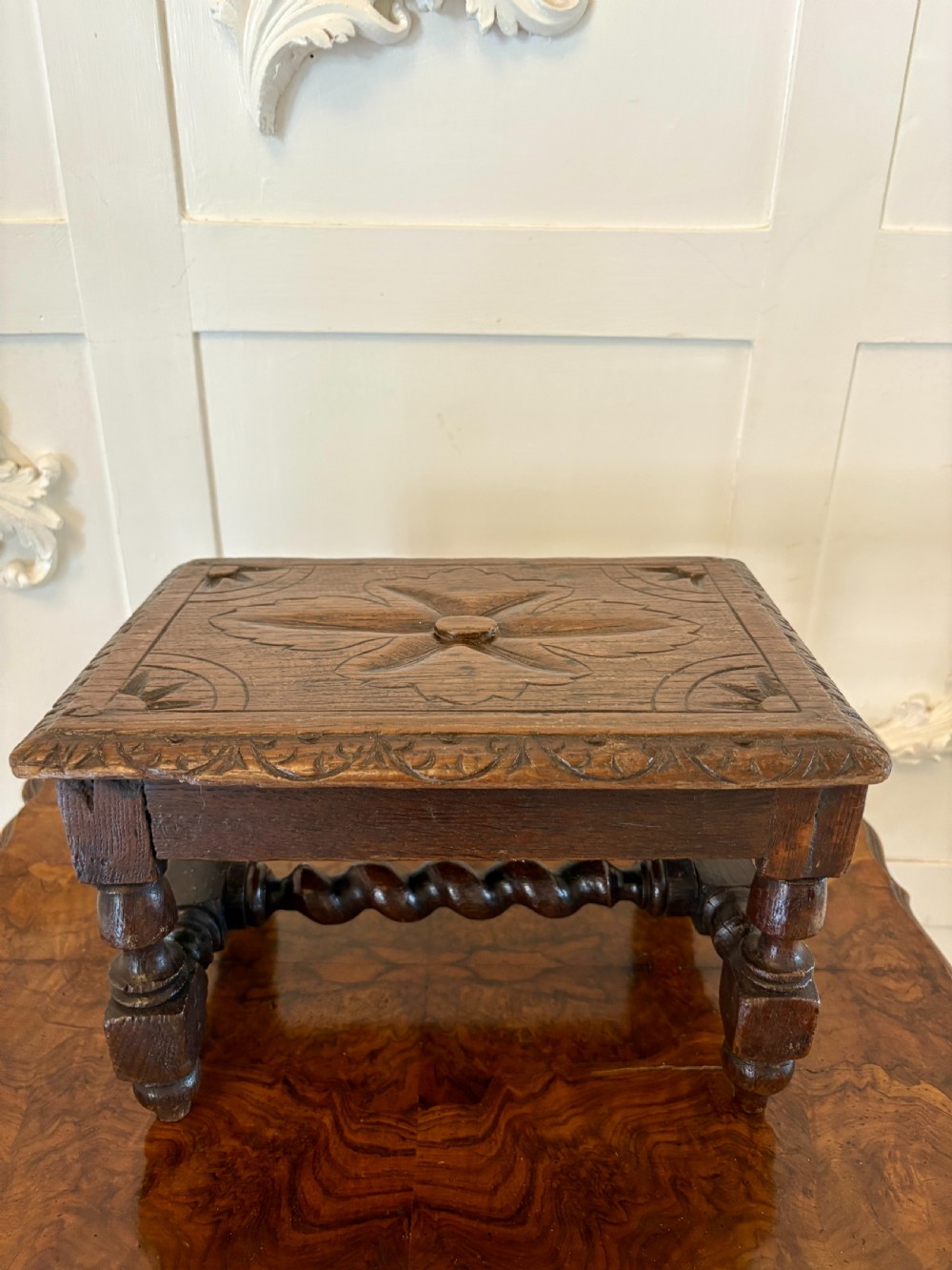 A dark carved oak miniature table with a central carved flower motif on top, carved frieze edges, turned legs, and a barley twist cross stretcher.