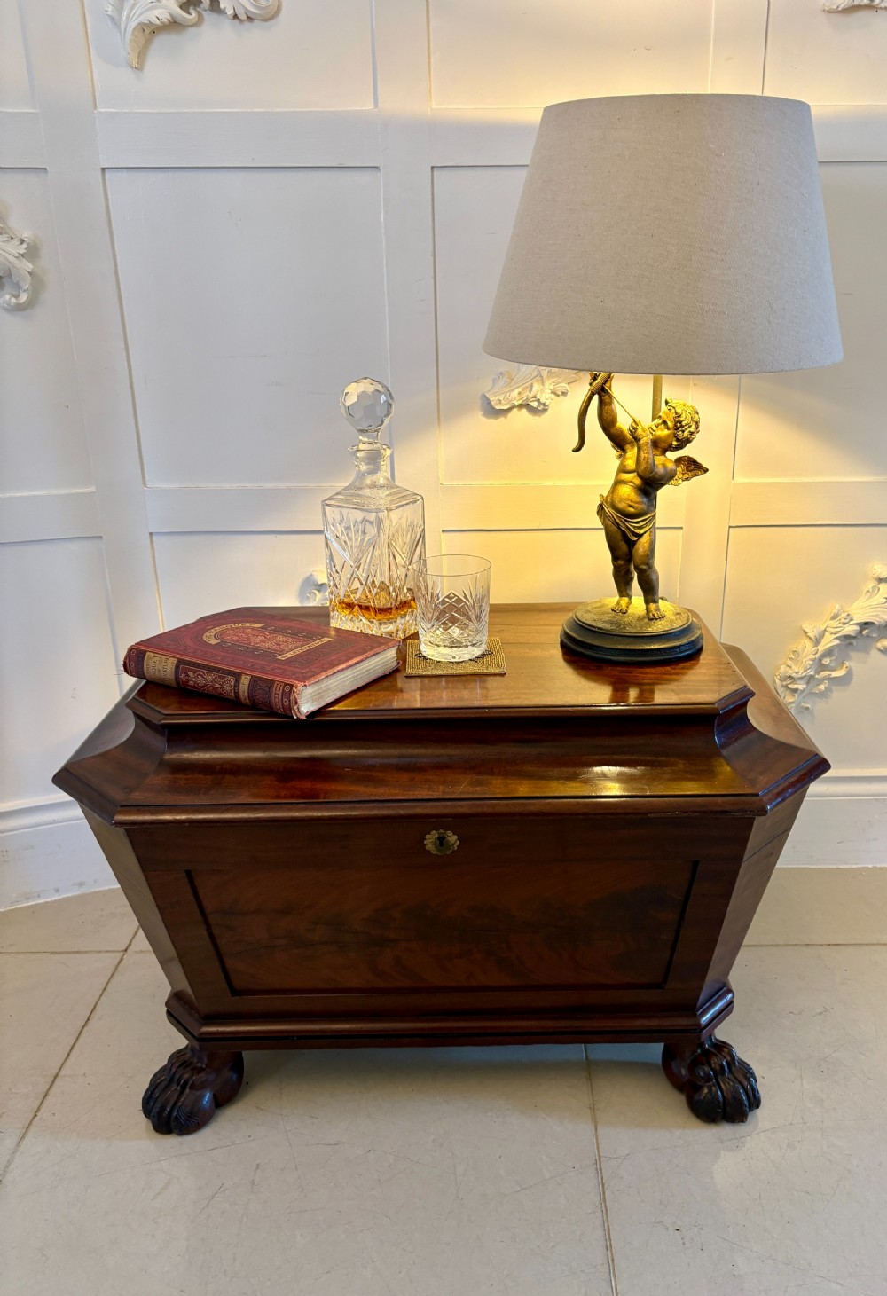 A Regency mahogany wine cooler shaped as a classical sarcophagus with figured wood panels, a central brass keyhole, and carved lion paw feet. View II