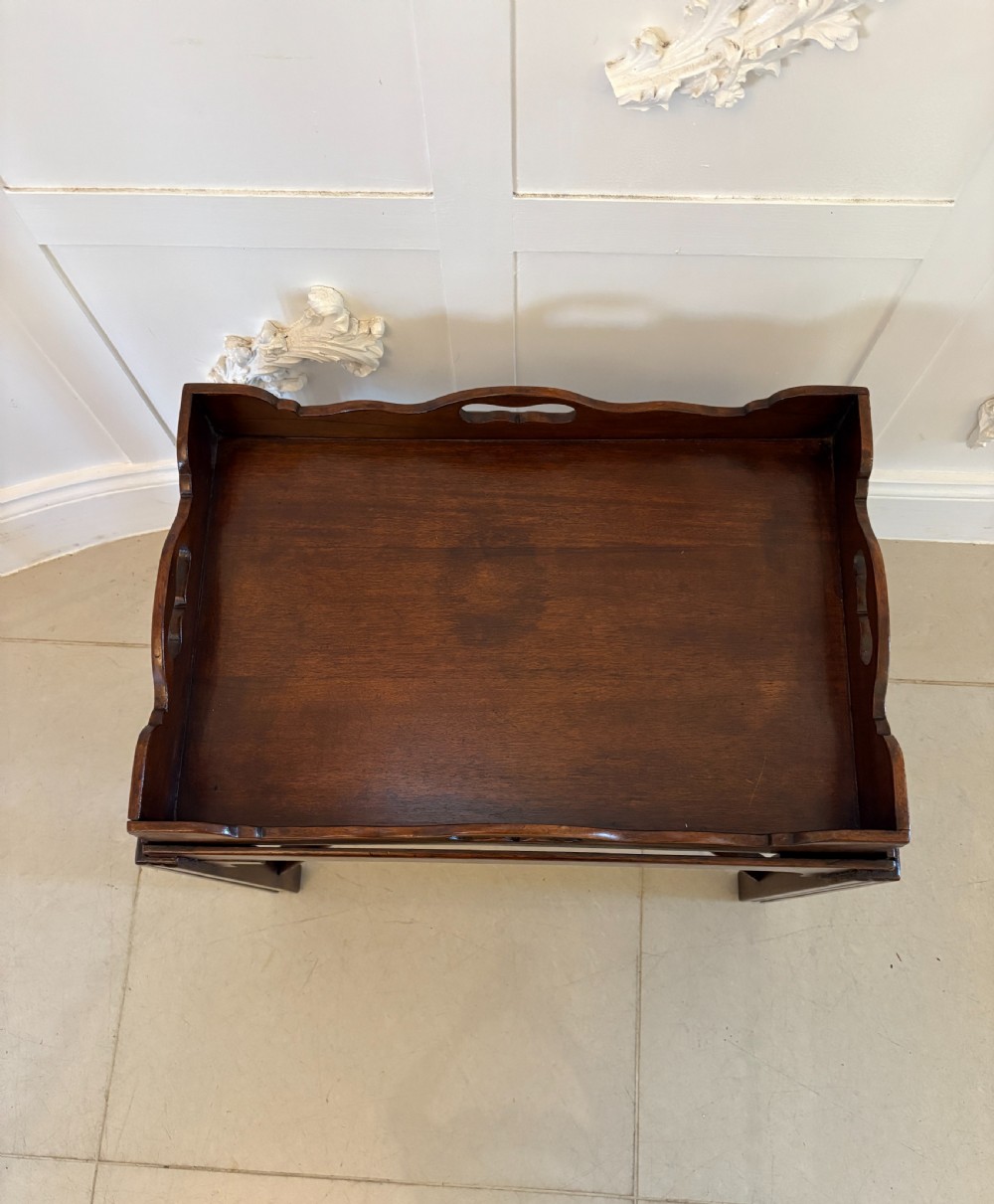 An Edwardian mahogany butlers tray featuring scalloped gallery sides with cut-out handles, resting on a matching wooden stand with reeded legs and cross-stretchers. View IV