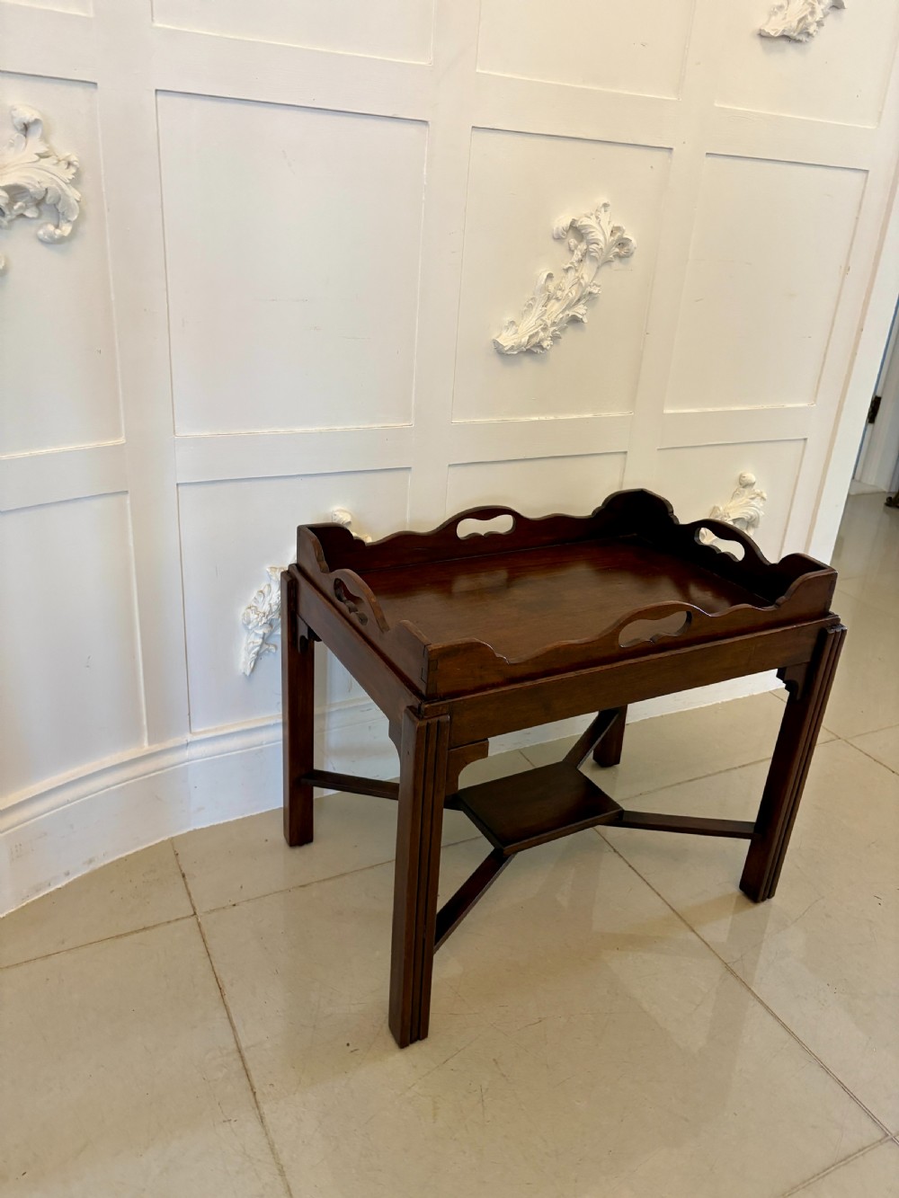 An Edwardian mahogany butlers tray featuring scalloped gallery sides with cut-out handles, resting on a matching wooden stand with reeded legs and cross-stretchers. View V