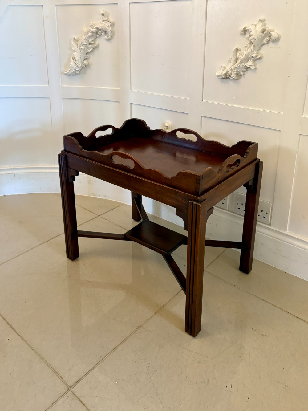 An Edwardian mahogany butlers tray featuring scalloped gallery sides with cut-out handles, resting on a matching wooden stand with reeded legs and cross-stretchers. View VI