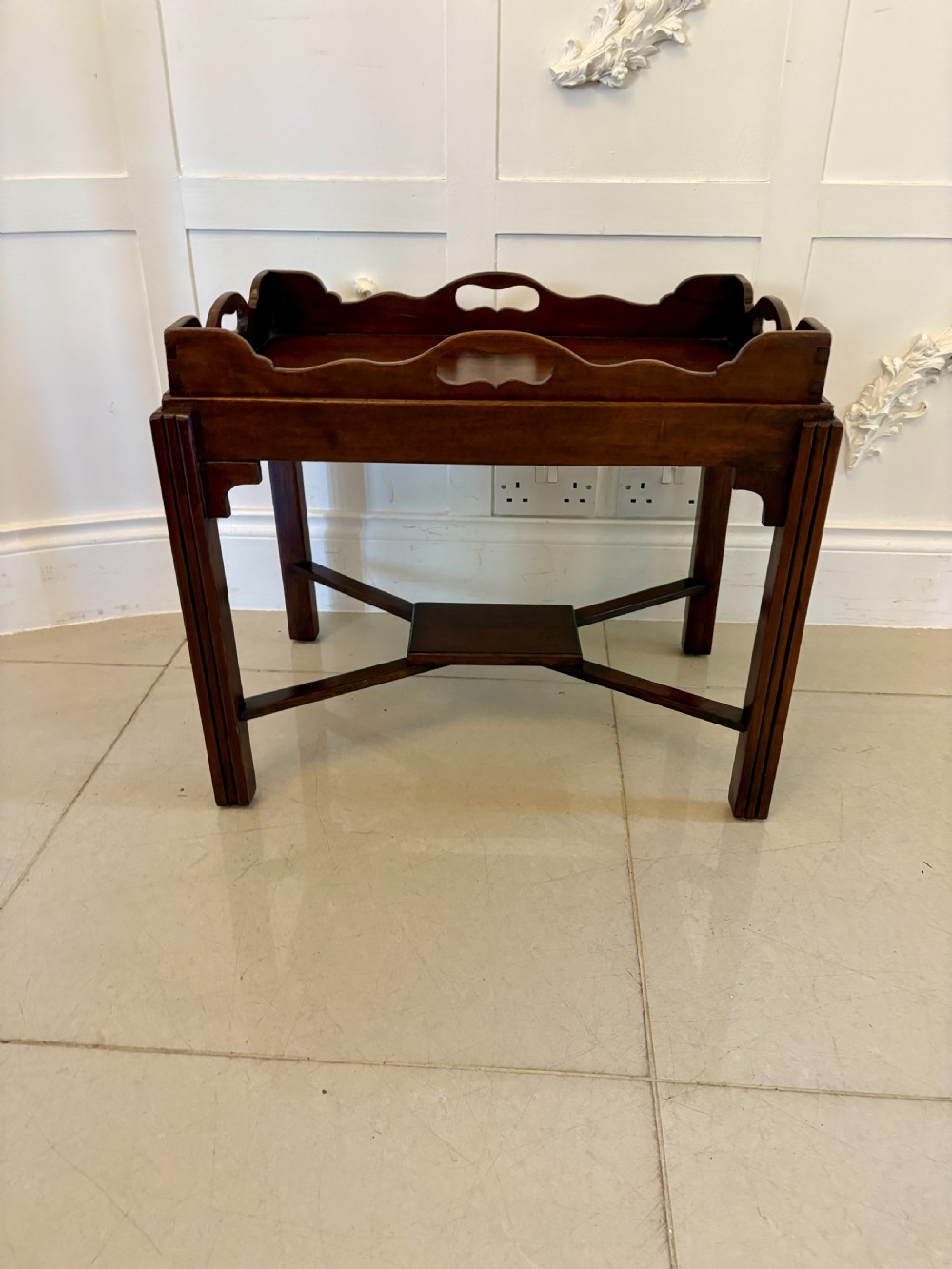 An Edwardian mahogany butlers tray featuring scalloped gallery sides with cut-out handles, resting on a matching wooden stand with reeded legs and cross-stretchers. View VII