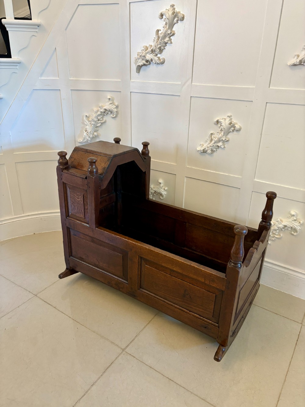 A dark oak panelled rocking crib featuring a rectangular frame, hooded end, turned corner finials, and curved base rockers.