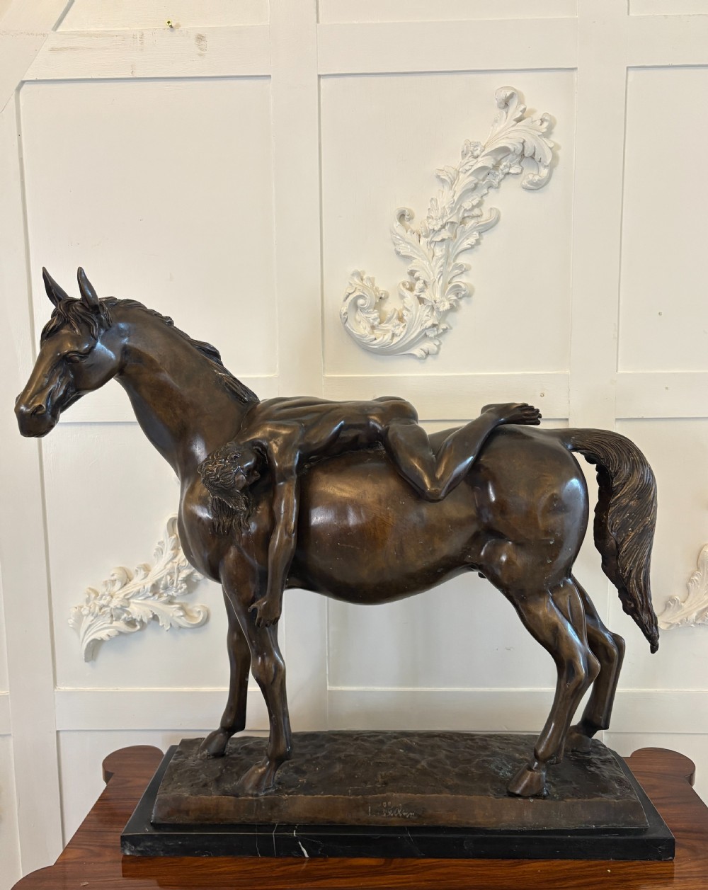 A dark brown bronze sculpture depicting a nude male figure draped across the back of a standing horse, mounted on a rectangular black marble base.