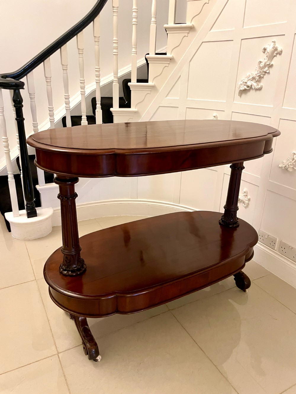 An oval dark mahogany two-tiered serving table featuring turned support columns, metal scroll handles, and carved feet with casters. View III