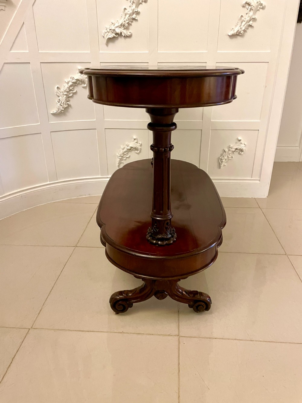 An oval dark mahogany two-tiered serving table featuring turned support columns, metal scroll handles, and carved feet with casters. View V