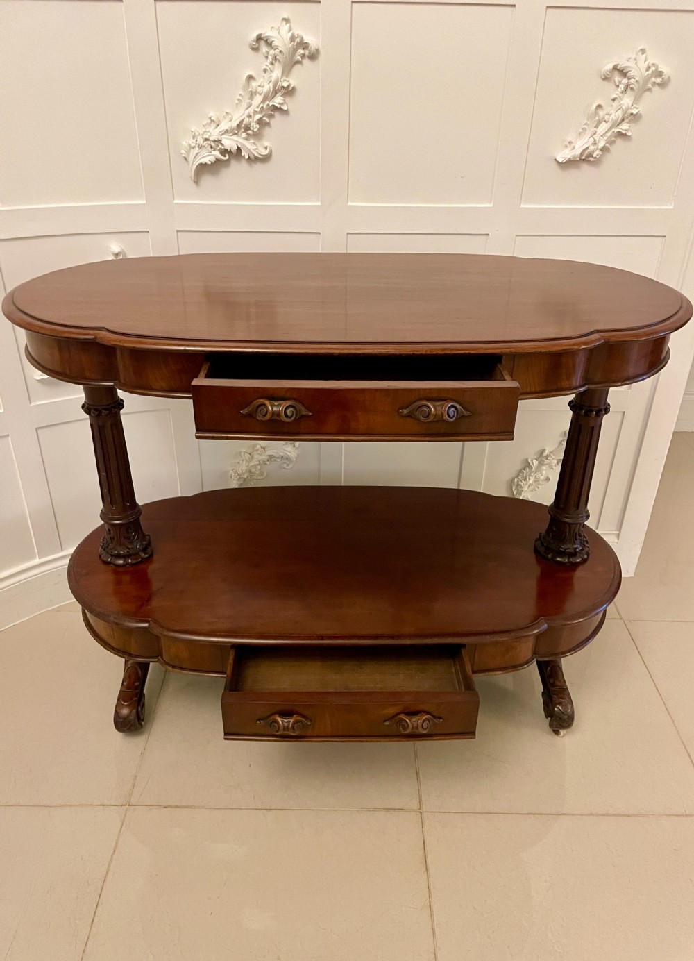 An oval dark mahogany two-tiered serving table featuring turned support columns, metal scroll handles, and carved feet with casters. View VII