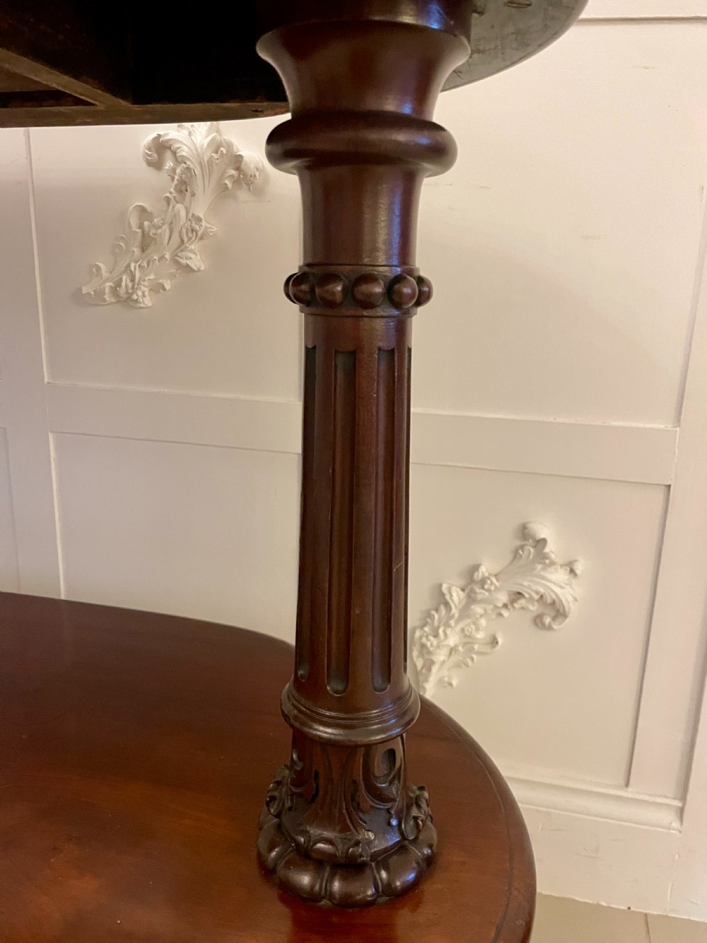 An oval dark mahogany two-tiered serving table featuring turned support columns, metal scroll handles, and carved feet with casters. View X
