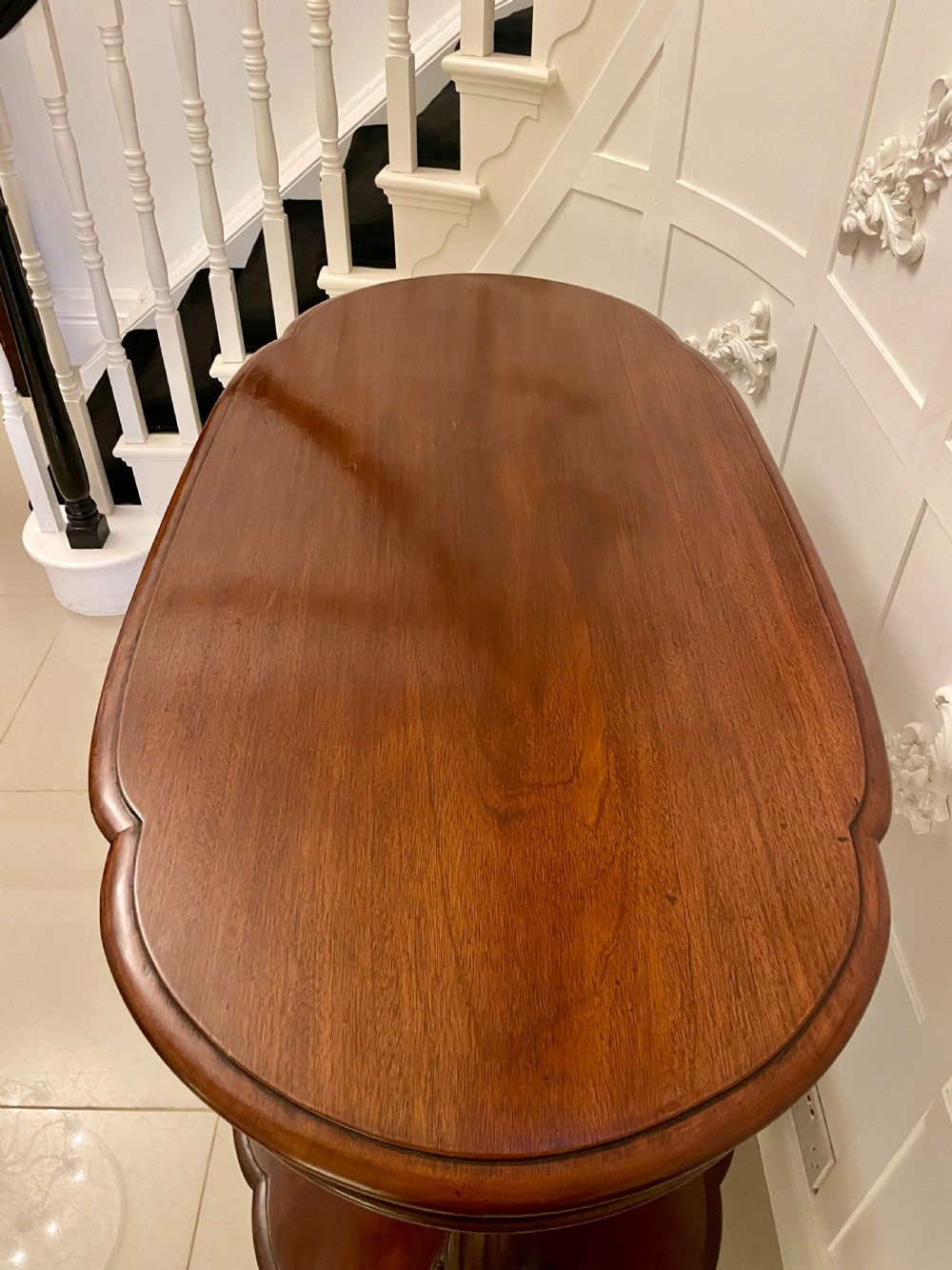 An oval dark mahogany two-tiered serving table featuring turned support columns, metal scroll handles, and carved feet with casters. View XII