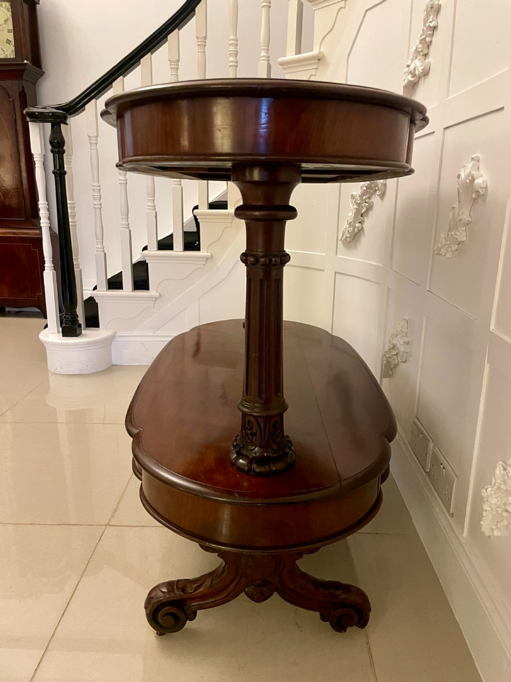 An oval dark mahogany two-tiered serving table featuring turned support columns, metal scroll handles, and carved feet with casters. View XIV