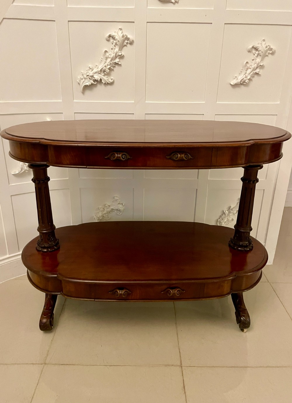 An oval dark mahogany two-tiered serving table featuring turned support columns, metal scroll handles, and carved feet with casters. View XV