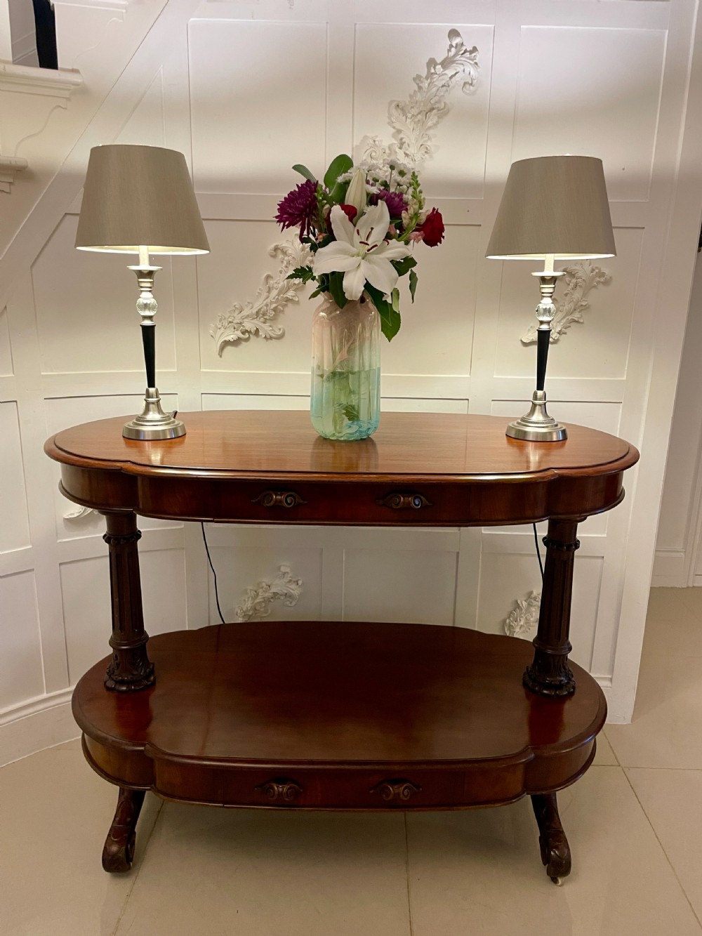 An oval dark mahogany two-tiered serving table featuring turned support columns, metal scroll handles, and carved feet with casters. View XVI