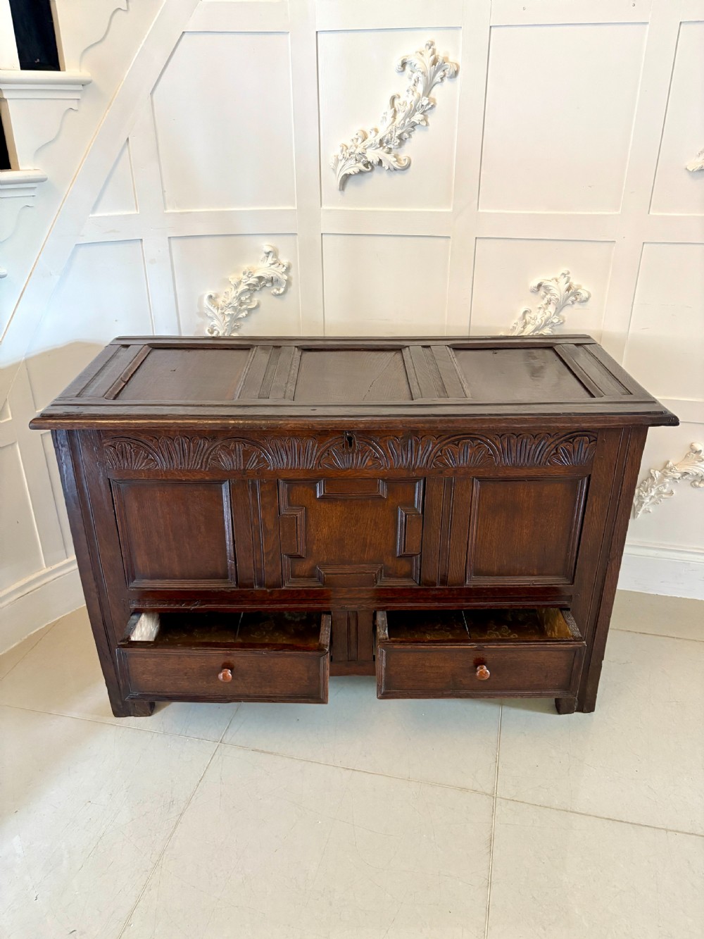 A seventeenth-century dark oak mule chest featuring a carved top frieze, geometric carved central panel, and two lower drawers with wooden turnings. View V