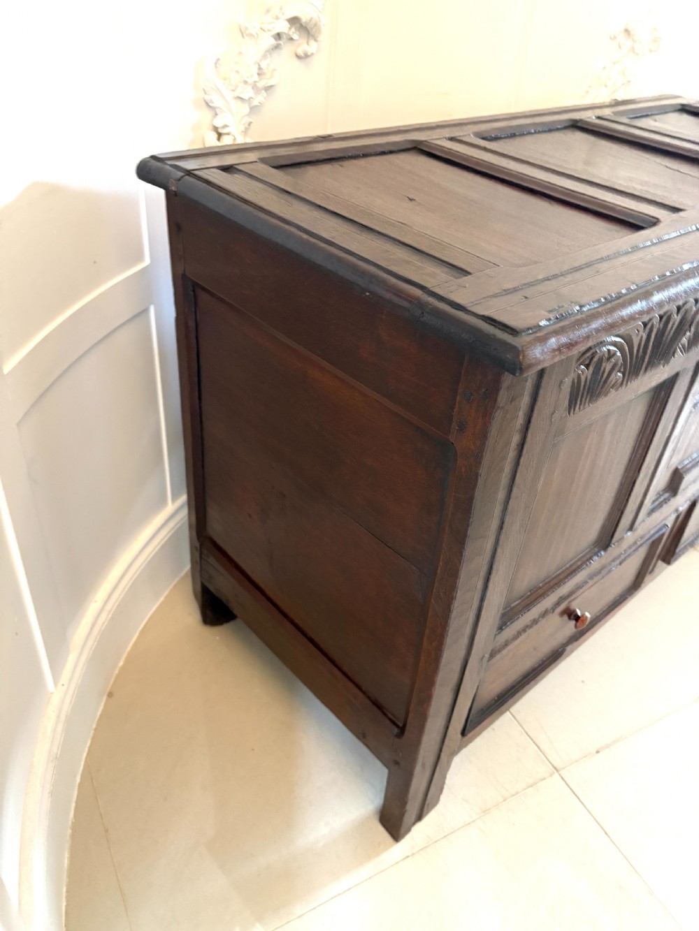 A seventeenth-century dark oak mule chest featuring a carved top frieze, geometric carved central panel, and two lower drawers with wooden turnings. View X