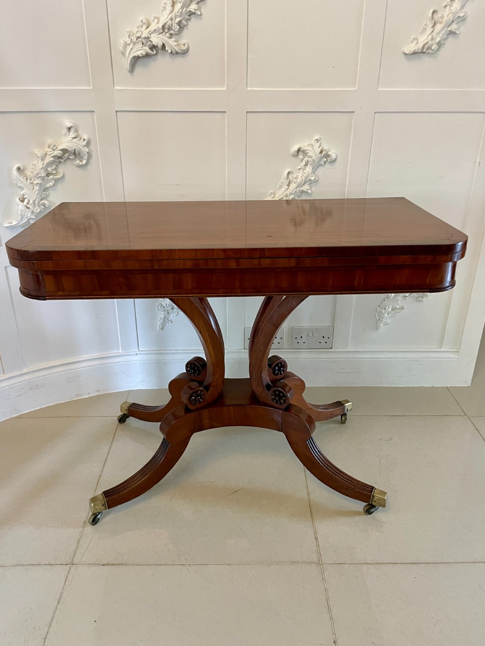 A Regency mahogany fold-over card table featuring a curved support pedestal, carved rosette scrolls, and four splayed legs fitted with brass castors.