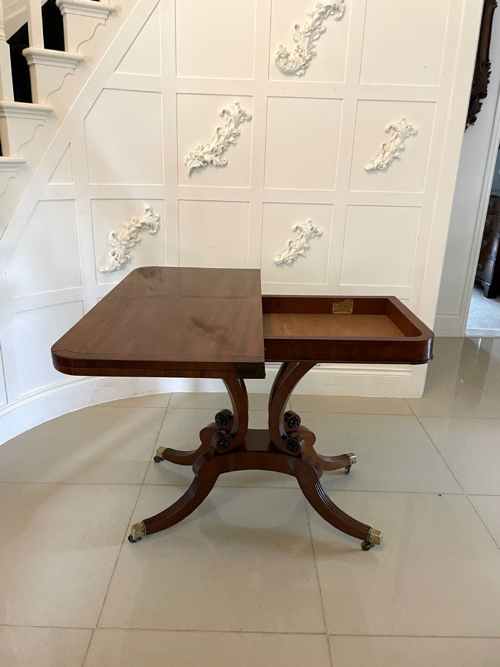 A Regency mahogany fold-over card table featuring a curved support pedestal, carved rosette scrolls, and four splayed legs fitted with brass castors. View III