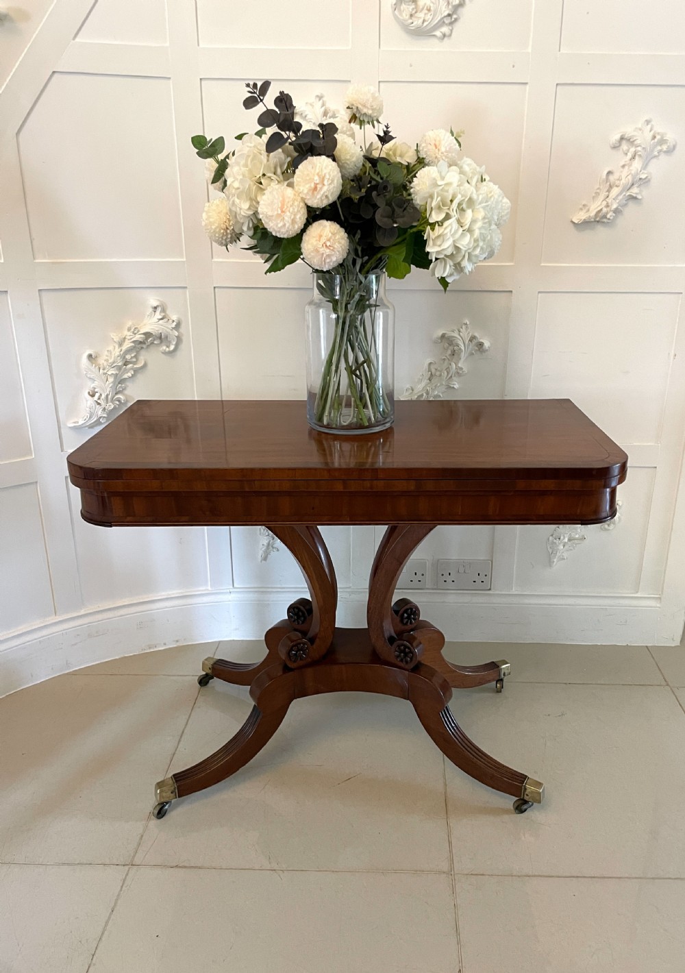 A Regency mahogany fold-over card table featuring a curved support pedestal, carved rosette scrolls, and four splayed legs fitted with brass castors. View V