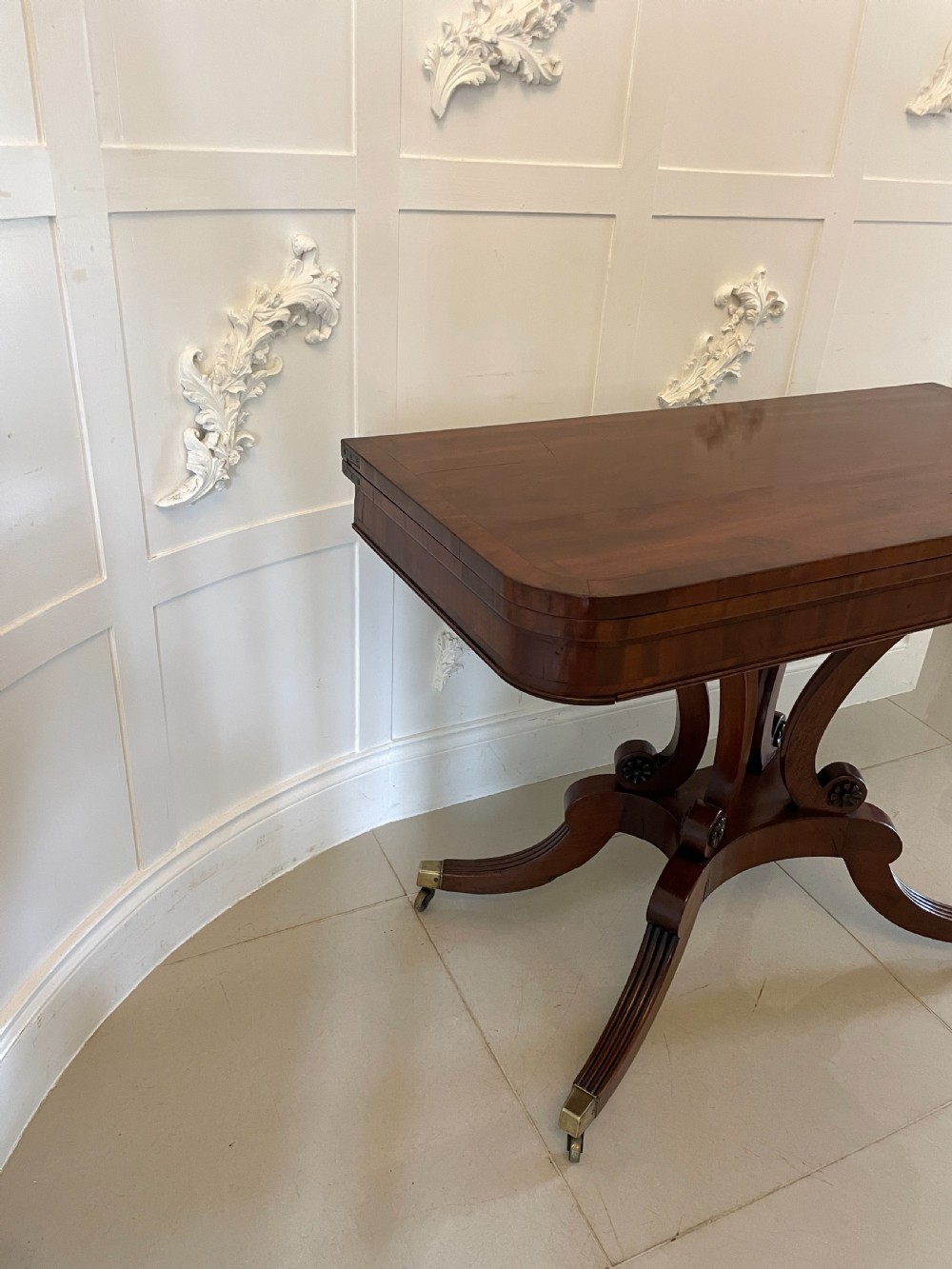 A Regency mahogany fold-over card table featuring a curved support pedestal, carved rosette scrolls, and four splayed legs fitted with brass castors. View XII