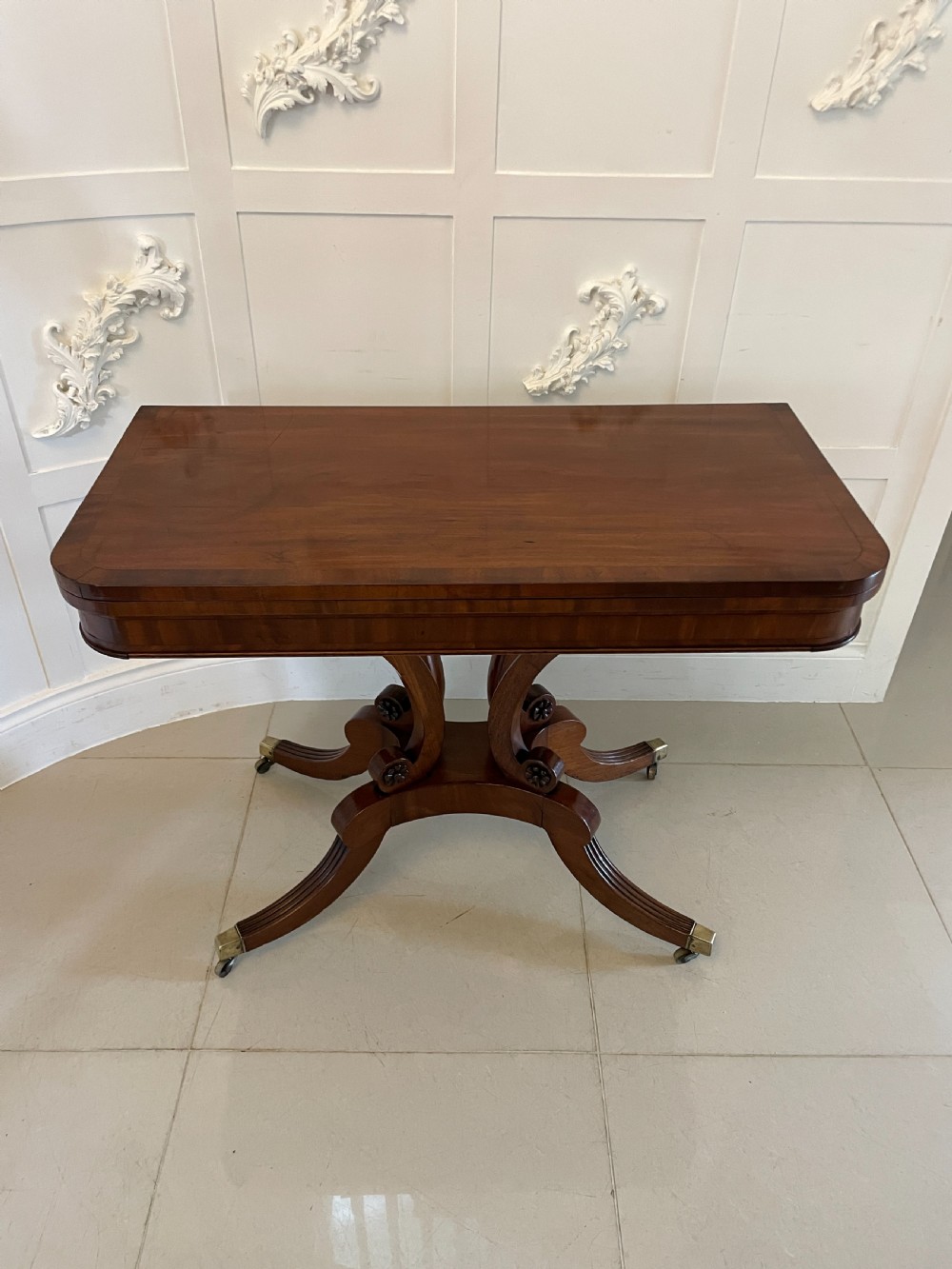 A Regency mahogany fold-over card table featuring a curved support pedestal, carved rosette scrolls, and four splayed legs fitted with brass castors. View XIII