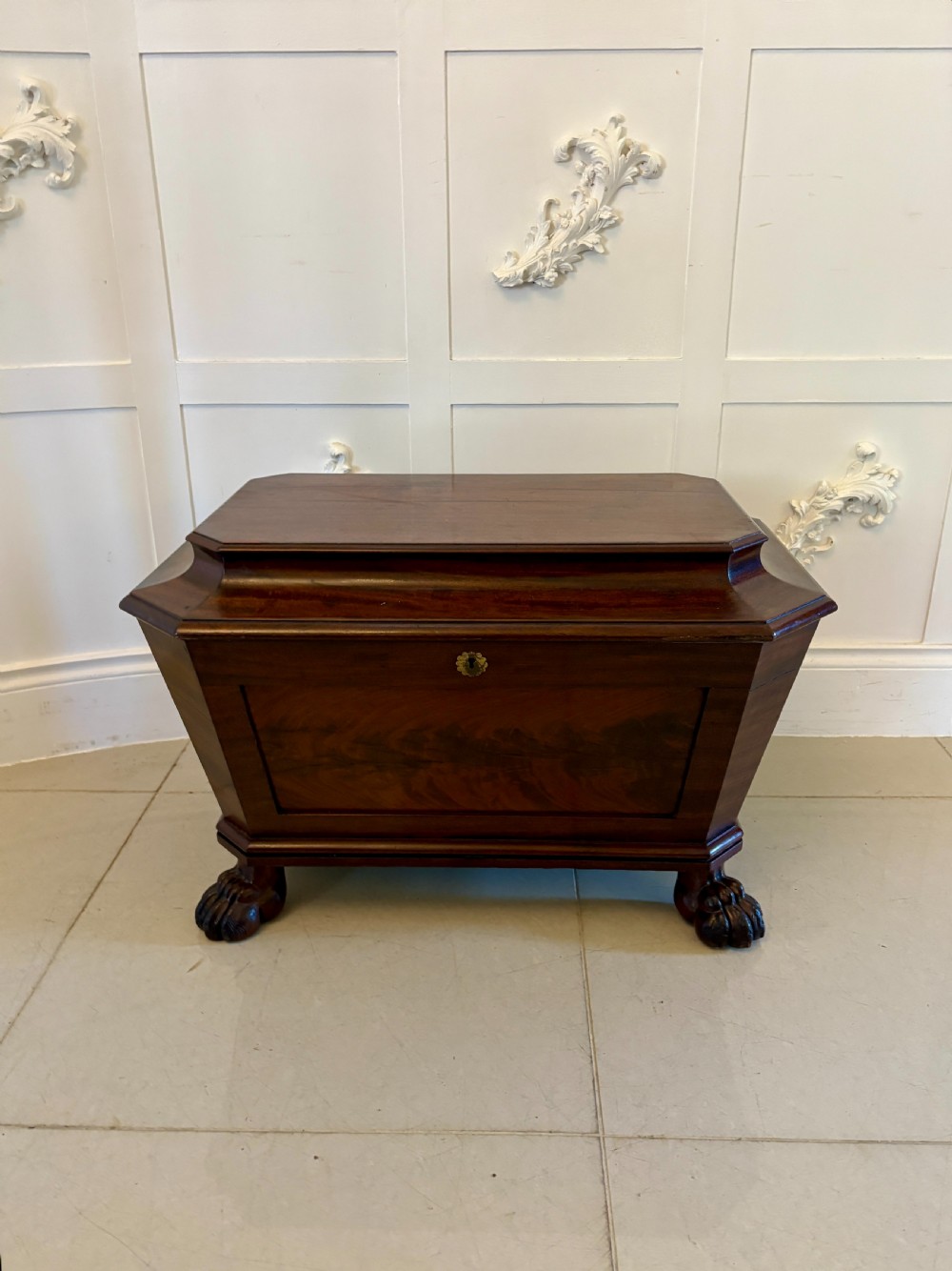 A Regency mahogany wine cooler shaped as a classical sarcophagus with figured wood panels, a central brass keyhole, and carved lion paw feet.