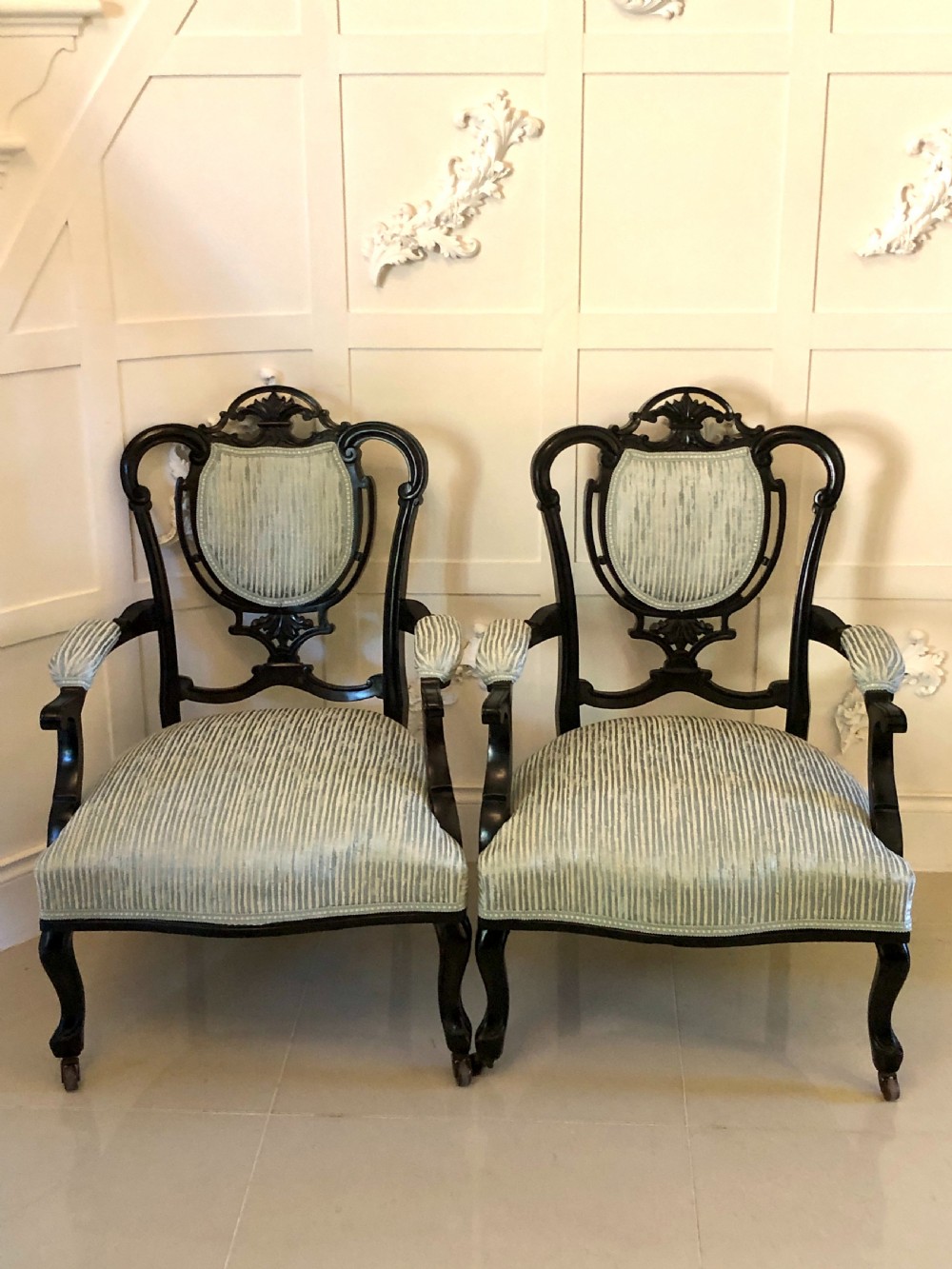 A pair of Victorian carved black lacquered open armchairs featuring oval padded backrests, curved padded arms, pale green striped fabric, and cabriole legs with brass castors.