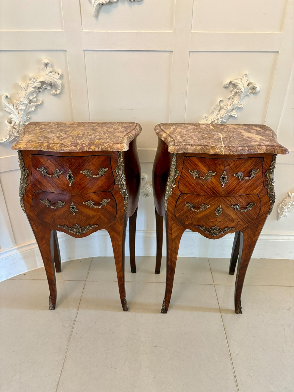 A pair of French Louis XV style kingwood bedside cabinets featuring shaped variegated marble tops, serpentine two-drawer fronts with marquetry inlay, gilt bronze mounts, and slender cabriole legs.