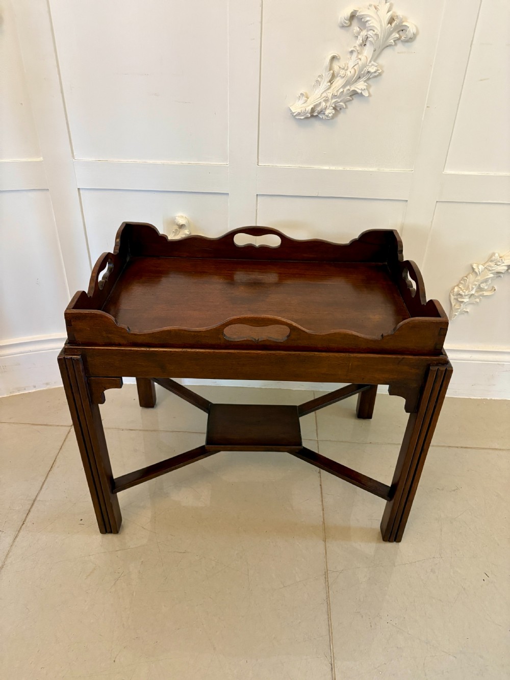 An Edwardian mahogany butlers tray featuring scalloped gallery sides with cut-out handles, resting on a matching wooden stand with reeded legs and cross-stretchers.