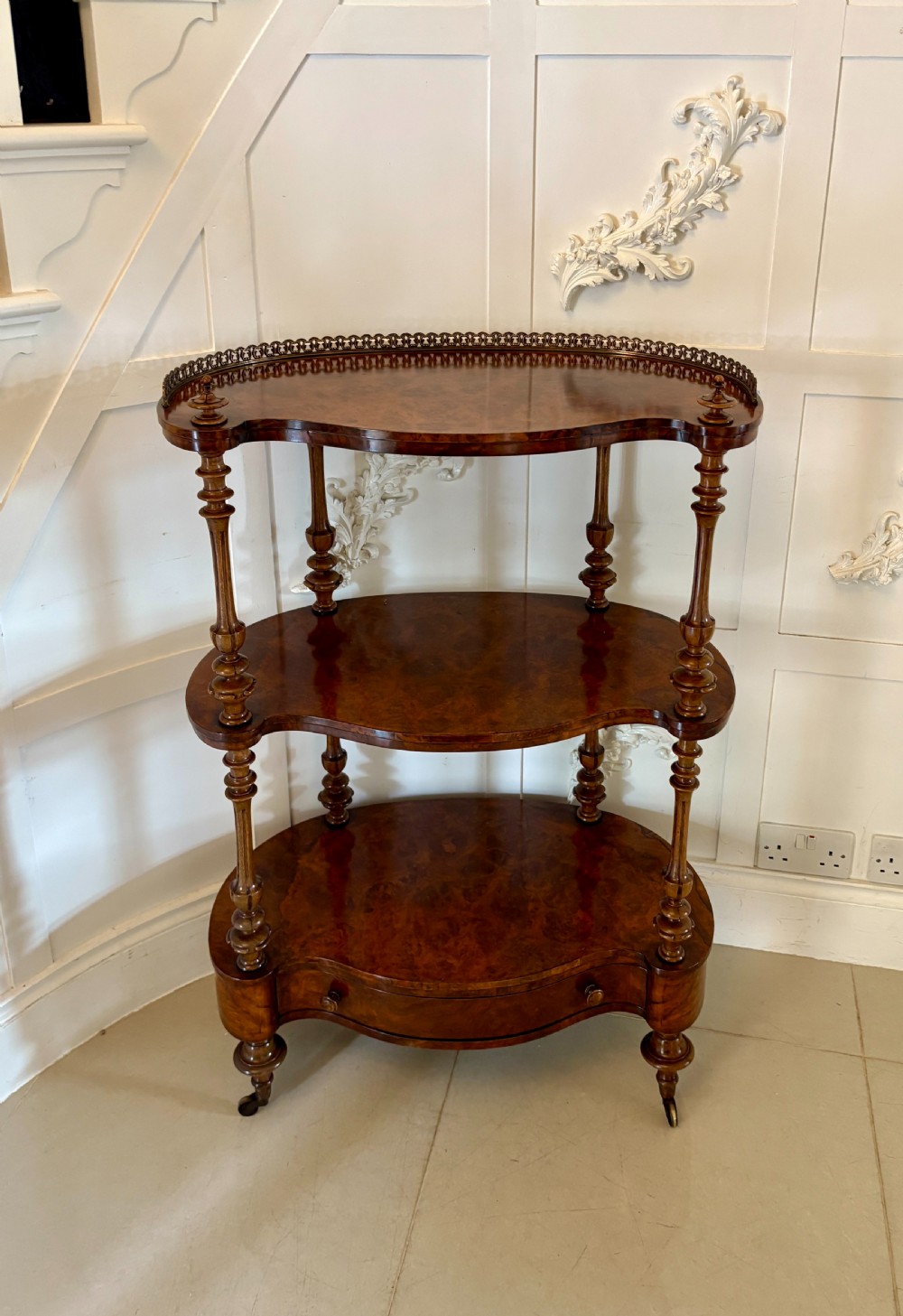 A Victorian three-tiered burr walnut whatnot featuring serpentine shelves, turned spindle supports, a pierced brass top gallery, and a base drawer on brass castors.