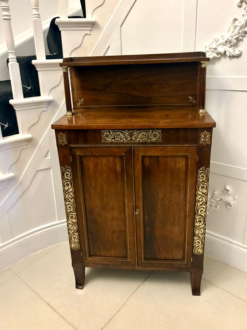 A Regency mahogany chiffonier featuring brass inlaid scroll motifs, a raised upper shelf supported by turned pillars, and a two-door lower cupboard.