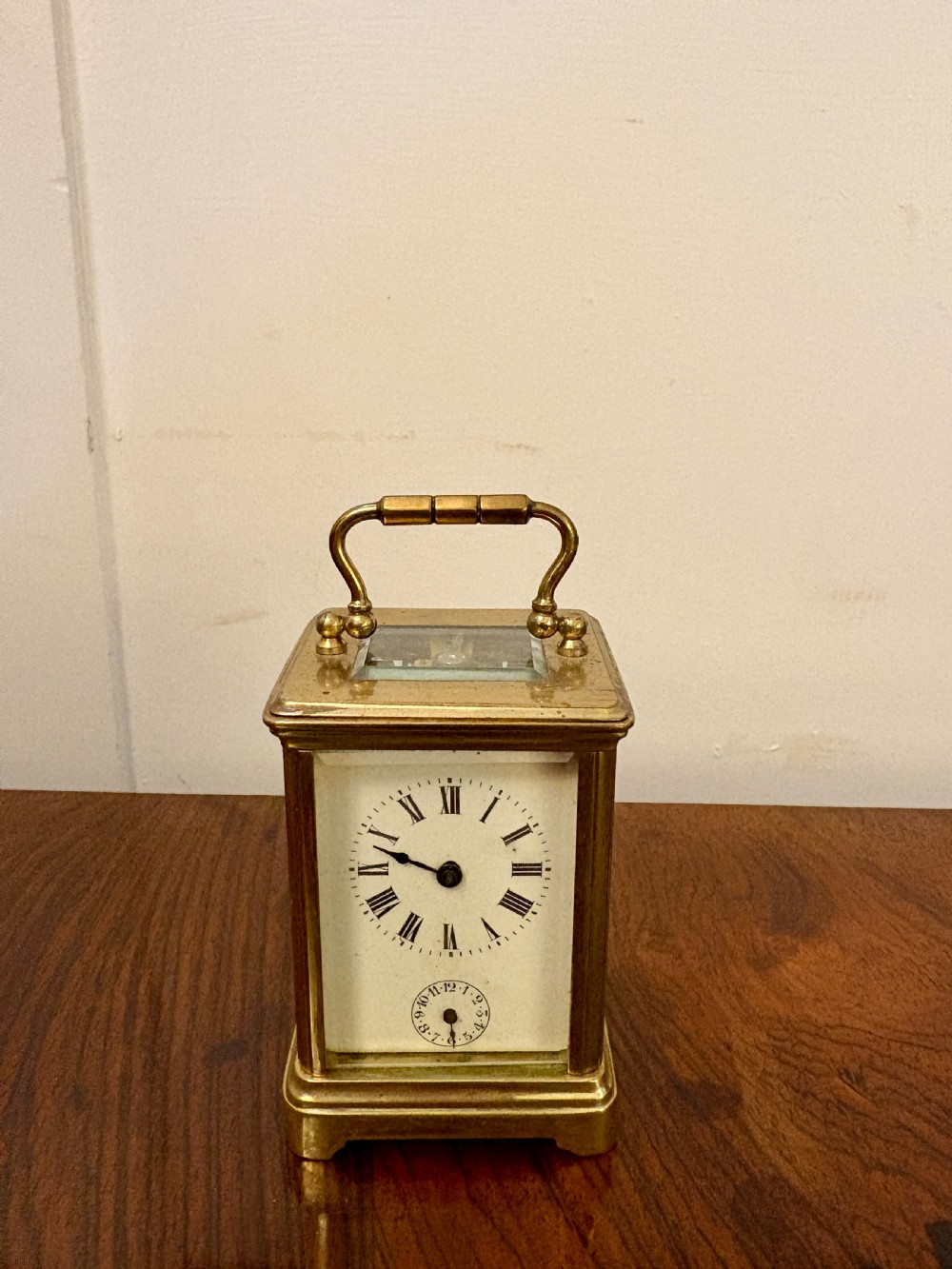 A miniature Victorian brass carriage clock featuring a white enamel dial with Roman numerals, an auxiliary alarm dial, glass panels, and a hinged carrying handle. View II