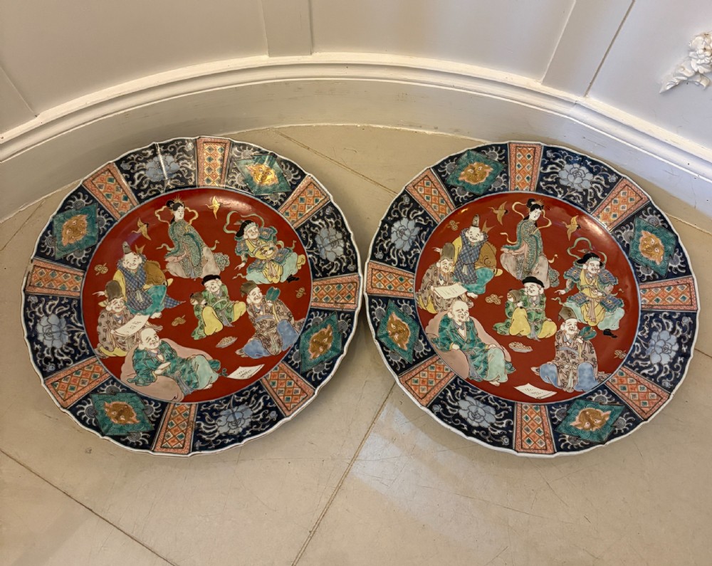 A pair of large circular Japanese Imari porcelain chargers featuring hand-painted figures of the Seven Lucky Gods framed by intricate cobalt blue and iron-red geometric borders.