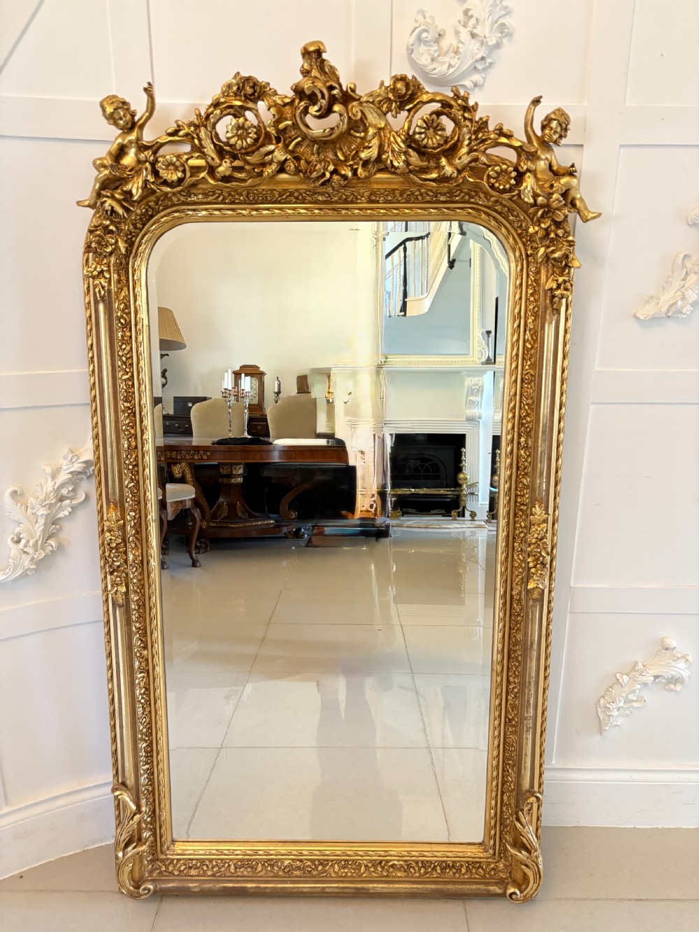 A large rectangular wall mirror featuring an ornate gilded frame decorated with carved floral scrolls and two cherub figures atop the crest.