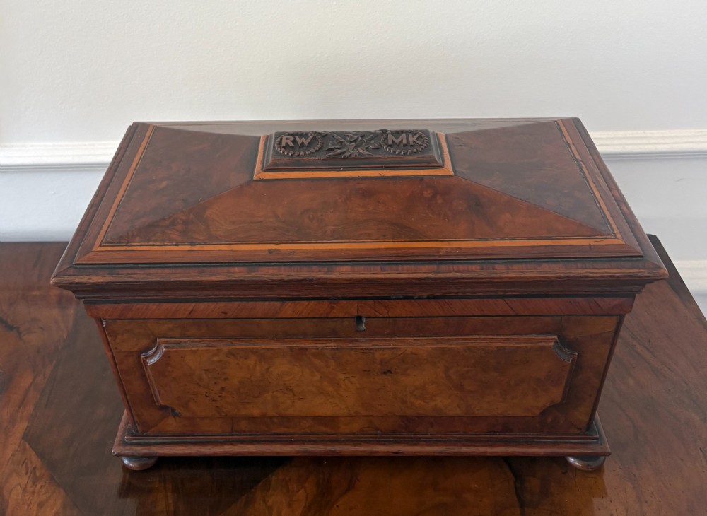A Victorian burr walnut sarcophagus-shaped tea caddy featuring a carved top plaque, stepped lid, keyhole escutcheon, and small bun feet. View V