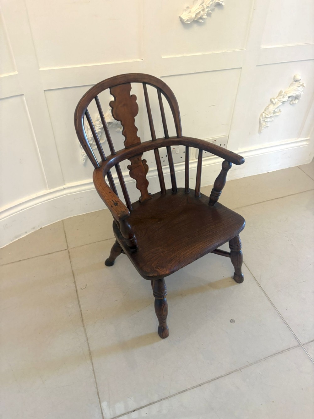 An antique Georgian childs Windsor armchair made of dark brown elm wood featuring a shaped back splat and turned legs.