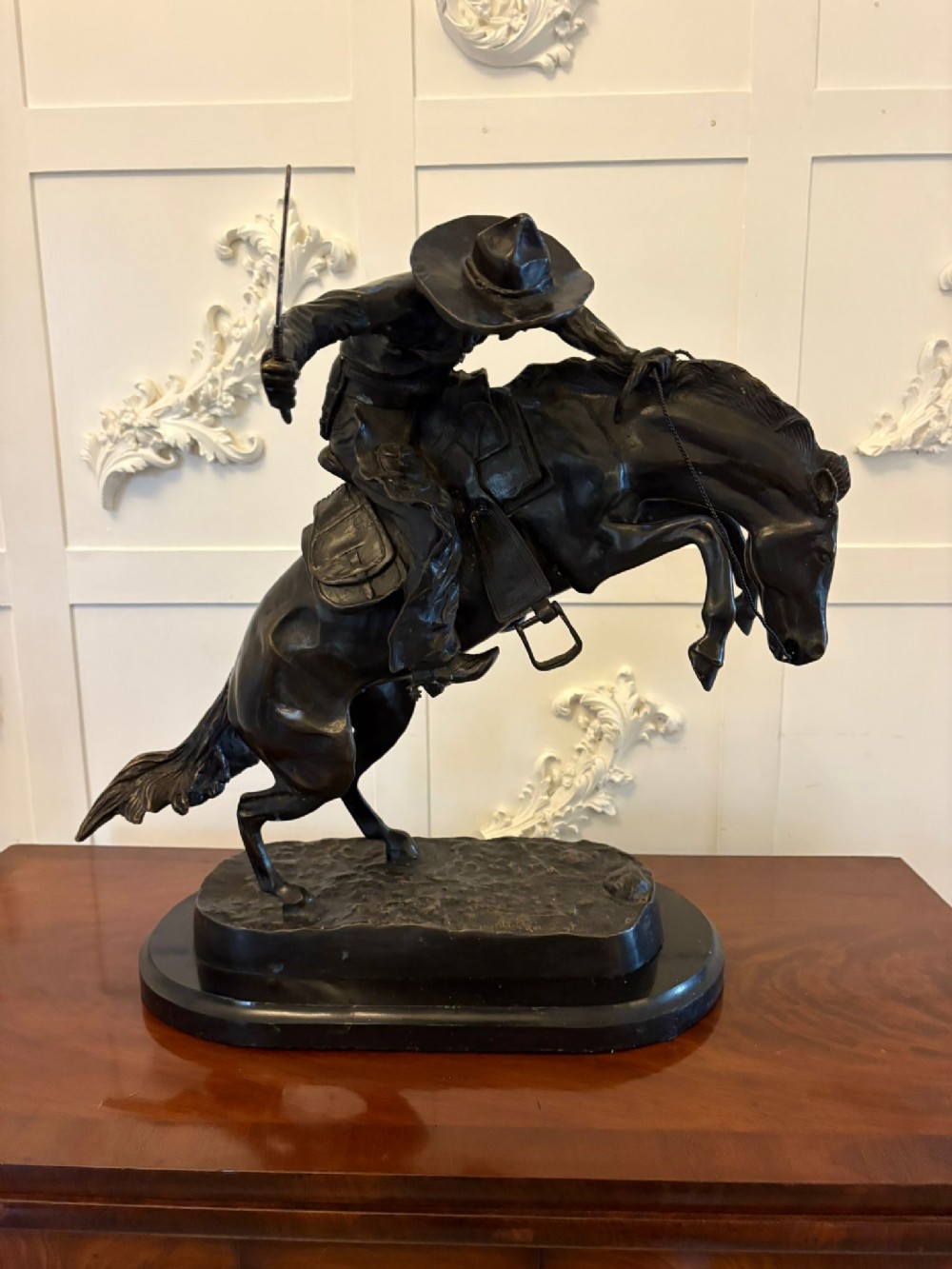 A dark patinated bronze sculpture depicting a cowboy wearing a hat atop a bucking horse on an oval base.