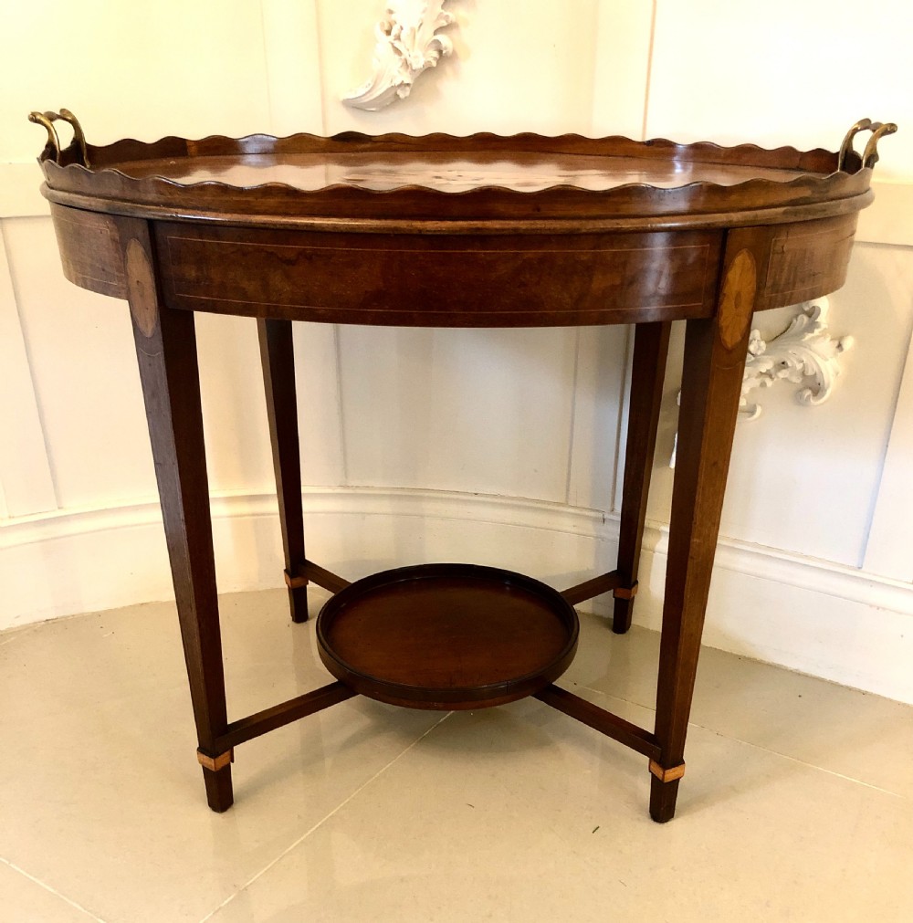 An Edwardian oval mahogany tray table featuring a scalloped gallery edge, brass side handles, satinwood inlay, and a lower circular shelf tier.